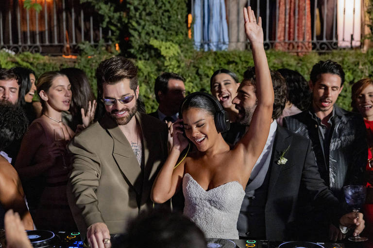 A imagem mostra uma festa animada com um DJ e uma mulher dançando. O DJ, um homem com cabelo curto e barba, usa óculos escuros e uma jaqueta clara, enquanto toca em uma mesa de som. A mulher, vestida com um vestido de noiva, usa fones de ouvido e levanta uma mão, sorrindo. Ao fundo, há várias pessoas dançando e se divertindo, com um ambiente festivo e decorado.