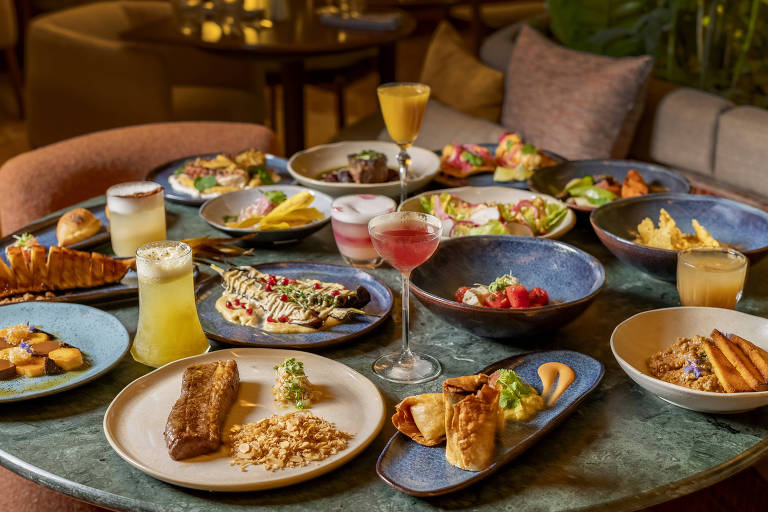 Uma mesa redonda com diversos pratos de comida dispostos. Há pratos com saladas, carnes, frutas, sobremesas e bebidas. Os pratos são de diferentes tamanhos e cores, com algumas bebidas em copos decorativos. O ambiente parece ser um restaurante elegante, com sofás ao fundo e uma iluminação suave.