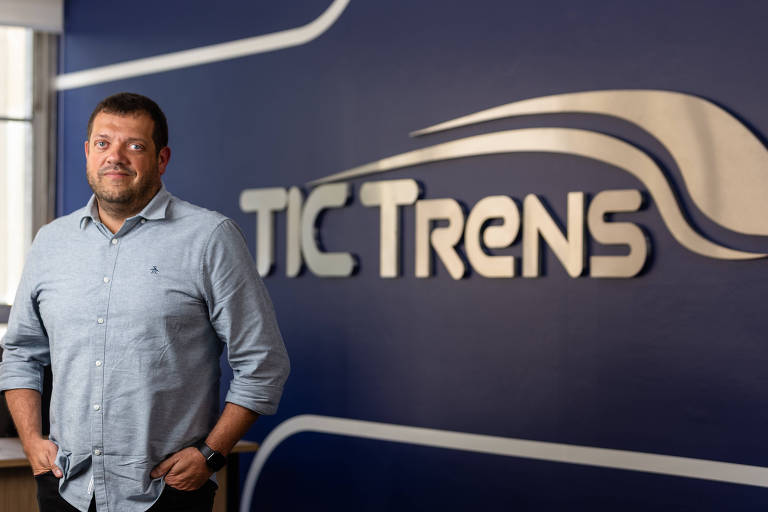 Um homem está posicionado à frente de uma parede com o logotipo da empresa 'TIC Trens'. Ele usa uma camisa de botão clara e está sorrindo levemente. O ambiente parece ser um escritório moderno, com uma decoração simples e profissional.