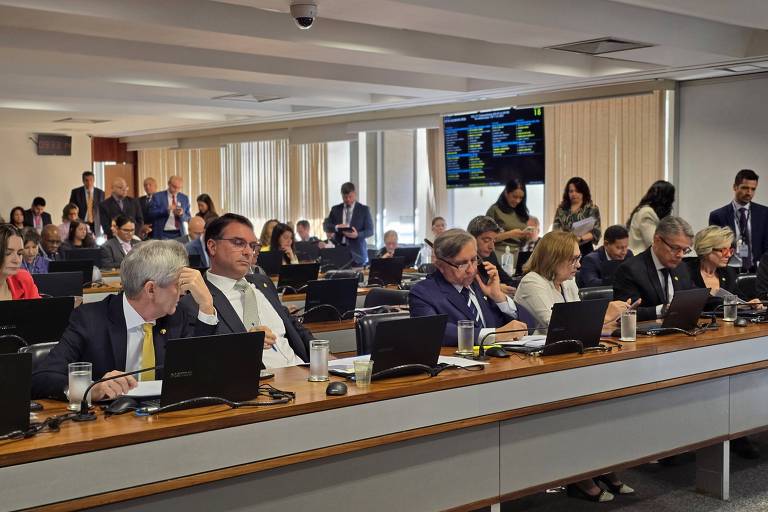 A imagem mostra uma sala de reuniões com várias pessoas sentadas em mesas, algumas usando laptops. Há um grupo de pessoas ao fundo, e uma tela de apresentação visível na parede, exibindo informações. A sala tem janelas grandes com luz natural entrando, e as pessoas parecem estar envolvidas em discussões ou trabalho.