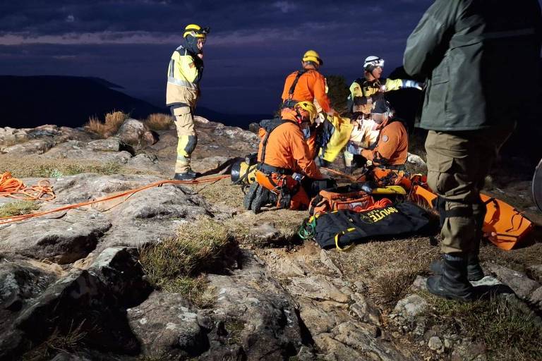 The image shows a rescue team in a mountainous area at night. Several team members are dressed in rescue uniforms, including helmets and reflective vests. They are gathered around a person who appears to be receiving treatment. Ropes and rescue equipment are scattered across the ground, and the lighting is low, with dark clouds in the background.