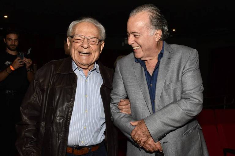 Na imagem, dois homens estão sorrindo e se divertindo em um evento. O homem à esquerda usa uma jaqueta de couro escura e uma camisa clara, enquanto o homem à direita veste um paletó cinza e uma camisa azul. Eles estão em um ambiente interno, com cadeiras vermelhas ao fundo e algumas luzes no teto.