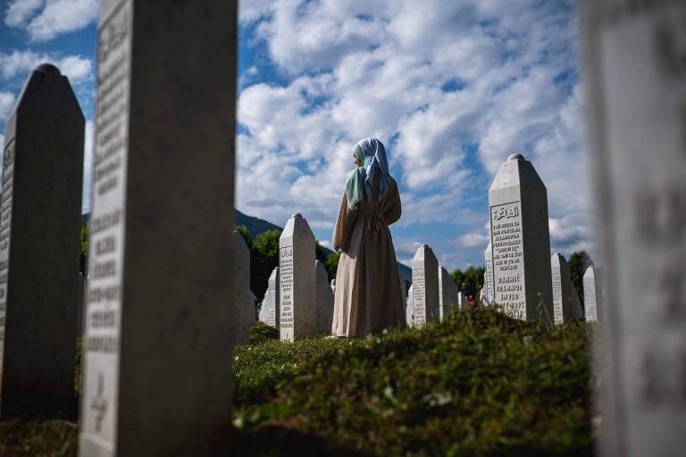 Uma mulher de costas, vestindo uma roupa longa e um lenço na cabeça, está em um cemitério cercada por lápides. O céu está parcialmente nublado, com nuvens brancas e um fundo azul. A grama é verde e bem cuidada, e as lápides são de pedra, algumas com inscrições visíveis.