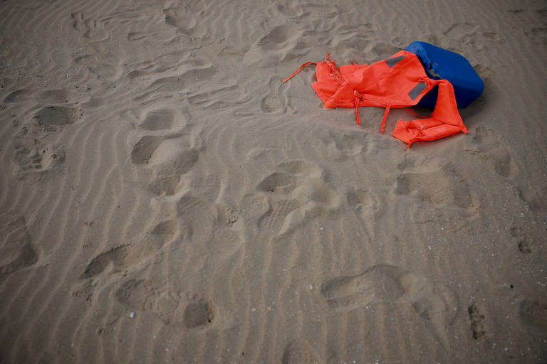 A imagem mostra uma área de areia com marcas de pegadas. No centro, há um colete salva-vidas laranja parcialmente dobrado e um objeto azul, possivelmente uma boia ou caixa, ao lado. O fundo é composto por areia com ondulações e mais pegadas visíveis.