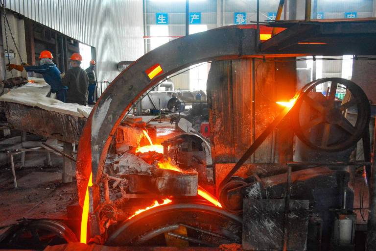 A imagem mostra um ambiente industrial de metalurgia, com um forno em funcionamento. O forno emite luz laranja intensa, indicando a presença de metal fundido. Ao fundo, há trabalhadores usando capacetes de segurança, observando o processo. O ambiente é predominantemente de metal e há equipamentos pesados visíveis.