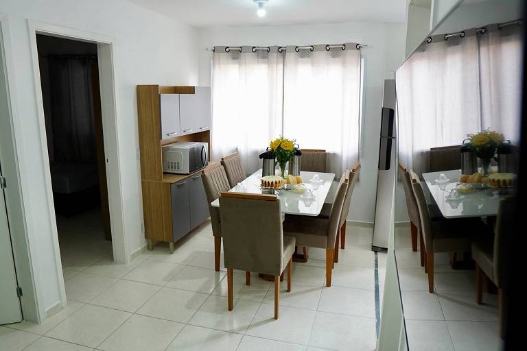 A imagem mostra uma cozinha integrada com uma sala de jantar. Há uma mesa retangular com cadeiras ao redor, e no centro da mesa, há um vaso com flores. Ao fundo, há um armário com um micro-ondas e prateleiras. As janelas têm cortinas claras, permitindo a entrada de luz natural. O piso é de cerâmica clara e as paredes são brancas.