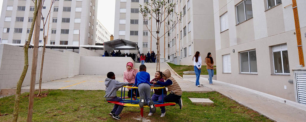 A imagem mostra uma área de lazer em um conjunto habitacional. No centro, crianças brincam em um equipamento de playground colorido, enquanto algumas árvores pequenas estão ao redor. Ao fundo, há prédios altos e modernos, com janelas e varandas. Algumas pessoas estão caminhando na área, e o céu está nublado.