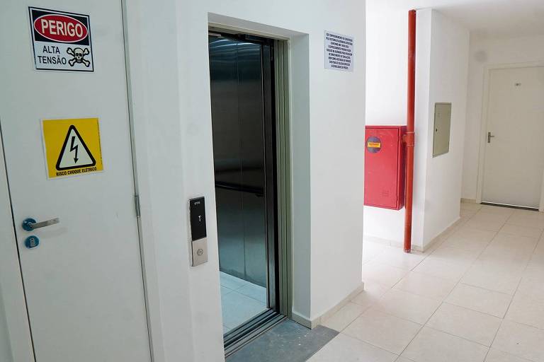 A imagem mostra um corredor com um elevador à esquerda. O elevador está aberto, revelando seu interior. Na parede ao lado do elevador, há um aviso de perigo com a inscrição 'PERIGO ALTA TENSÃO' e um símbolo de eletricidade. Também há um sinal de alerta amarelo com um símbolo de raio. À direita, há um extintor de incêndio vermelho e uma porta fechada. O piso é de cerâmica clara e as paredes são brancas.