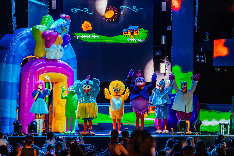 Um grupo de personagens coloridos em trajes de fantasia se apresenta em um palco. Eles estão em frente a um cenário com um fundo colorido que inclui um sol, uma casa e um campo verde. Os personagens estão fazendo gestos de saudação, enquanto uma estrutura inflável em forma de arco-íris está ao fundo. A plateia, composta por crianças e adultos, observa a apresentação.