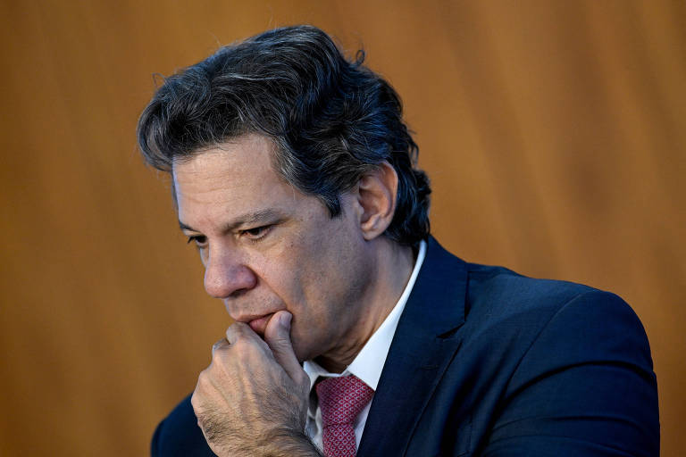 Um homem com cabelo castanho e ondulado, vestindo um terno escuro e uma gravata vermelha, está com a mão no queixo, olhando para baixo em um ambiente com fundo de madeira. Sua expressão é de reflexão ou preocupação