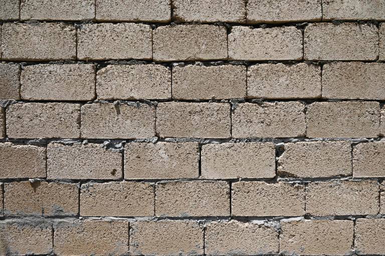 A imagem mostra uma parede feita de tijolos de cor clara, com uma textura rugosa. Os tijolos estão dispostos em fileiras horizontais, e algumas áreas apresentam desgaste e pequenas fissuras. A superfície parece ser de um material de construção comum, com uma aparência um pouco desgastada.