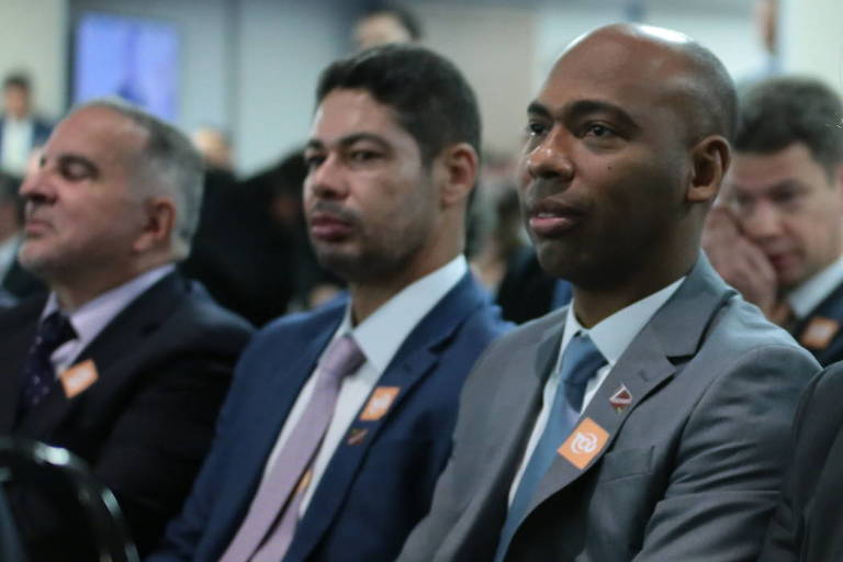 Um grupo de homens está sentado em um evento, todos vestindo trajes formais. Eles parecem estar prestando atenção ao que está acontecendo à sua frente. Alguns deles usam crachás de identificação. O ambiente é bem iluminado e há outras pessoas ao fundo, indicando que é um evento com público.
