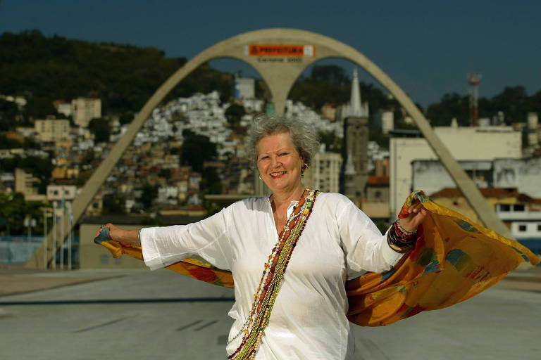A smiling woman stands in the foreground, wearing a white blouse and holding a colorful cloth. In the background, there's a large arch marked 'Ponte Estaiada' (Stayed Bridge) and a view of a city with houses stacked on a hill. The sky is clear, and the scene appears to be daytime.