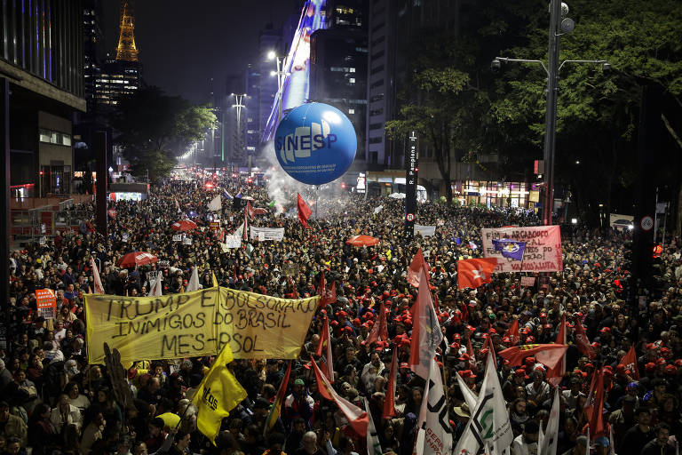 A imagem mostra uma grande multidão reunida em uma manifestação noturna em uma rua de São Paulo. Muitas pessoas seguram bandeiras vermelhas e cartazes. Um dos cartazes é amarelo e contém a frase 'TRUMP E BOLSONARO INIMIGOS DO BRASIL'. No fundo, há um grande balão azul com a inscrição 'NESPT'. Edifícios altos e árvores estão visíveis ao redor, e há iluminação urbana ao fundo.