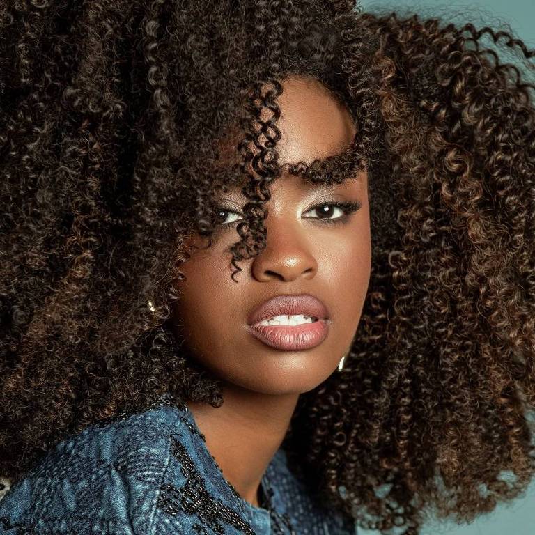 A imagem mostra uma mulher com cabelo cacheado e volumoso, que possui uma coloração castanho claro com mechas mais escuras. Ela está olhando diretamente para a câmera, com uma expressão confiante. A maquiagem é suave, destacando os lábios em um tom nude. A mulher usa uma blusa azul com um padrão texturizado. O fundo da imagem é de uma cor azul clara, criando um contraste com o cabelo e a roupa.