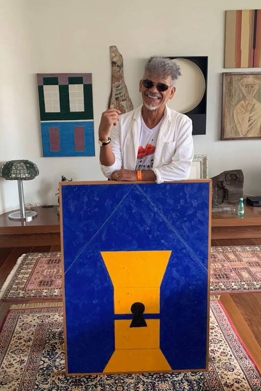 Uma pessoa está em pé segurando um objeto em uma sala de estar. O fundo apresenta uma mesa de madeira com uma lâmpada, um sofá e uma televisão. As paredes têm várias obras de arte, incluindo quadros com padrões e cores variadas. O piso é de madeira e há um tapete decorativo no chão.