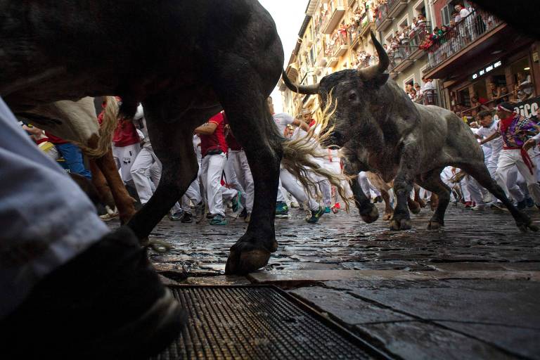 A imagem mostra uma cena de uma corrida de touros, com touros correndo em uma rua pavimentada. Pessoas vestidas de branco e vermelho estão ao redor, algumas correndo e outras observando. O foco está em um touro que parece estar se movendo rapidamente, enquanto as patas de um homem são visíveis em primeiro plano. O ambiente é urbano, com edifícios ao fundo e pessoas nas janelas.