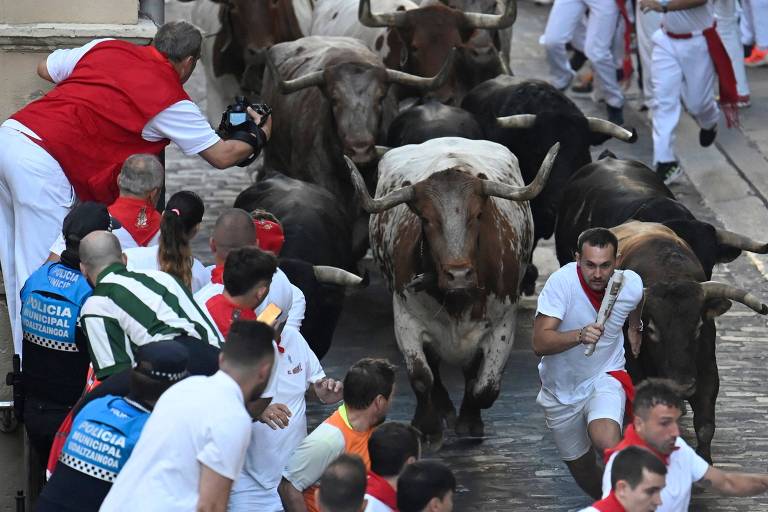 A imagem mostra um grupo de touros correndo em uma rua estreita, enquanto várias pessoas vestidas de branco e vermelho tentam se afastar. Algumas pessoas estão correndo em direção aos touros, enquanto outras estão se protegendo atrás de barreiras. Há também policiais presentes na cena, monitorando a situação.