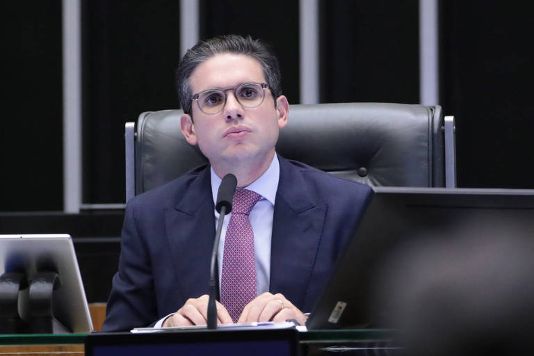 Um homem com óculos e cabelo liso, vestindo um terno escuro e uma gravata roxa, está sentado em uma cadeira de mesa em um ambiente legislativo. Ele parece estar ouvindo ou pensando, com uma expressão séria. À sua frente, há um microfone e um tablet. O fundo é composto por painéis verticais em cores escuras.