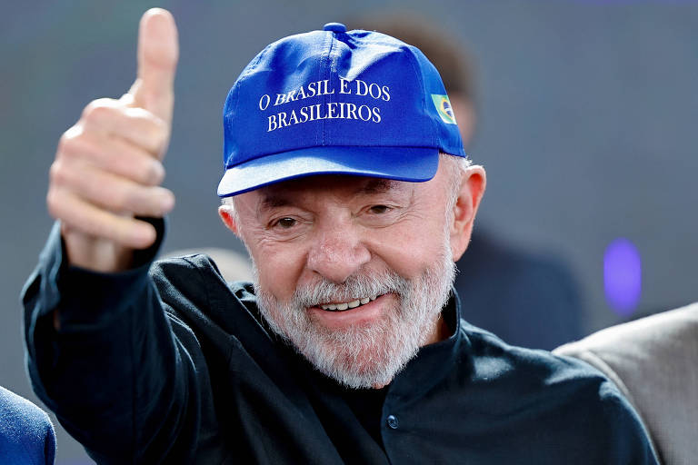 Um homem sorridente, com barba e cabelo grisalho, está fazendo um gesto de positivo com a mão. Ele usa um boné azul com a frase "O BRASIL É DOS BRASILEIROS" e uma bandeira do Brasil. O fundo é desfocado, sugerindo um evento ou uma cerimônia.