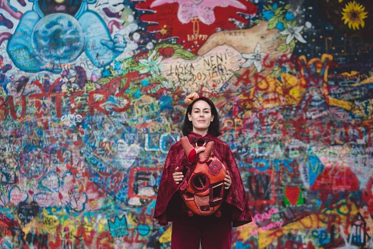 A imagem mostra uma mulher sorridente em pé diante de um mural vibrante e colorido, coberto de grafites e desenhos. Ela está vestindo uma capa vermelha e segurando uma escultura de coração, que parece ser feita de material macio. O fundo é uma mistura de cores e formas, com várias palavras e desenhos visíveis, criando um ambiente artístico e expressivo.