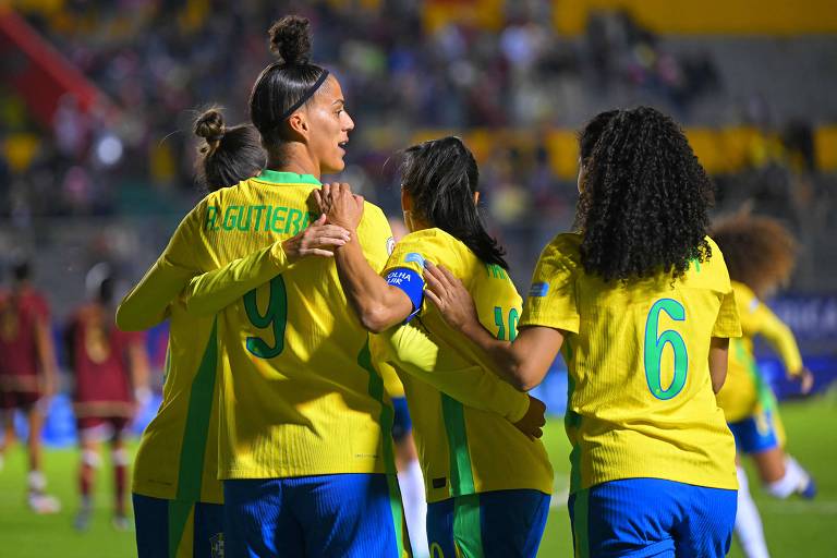 Um grupo de jogadoras de futebol vestindo uniformes amarelos e azuis do Brasil se abraçam em um momento de celebração. Uma jogadora, com o número 9 nas costas, está no centro da imagem, enquanto outras jogadoras a cercam. O fundo mostra um estádio com torcedores.