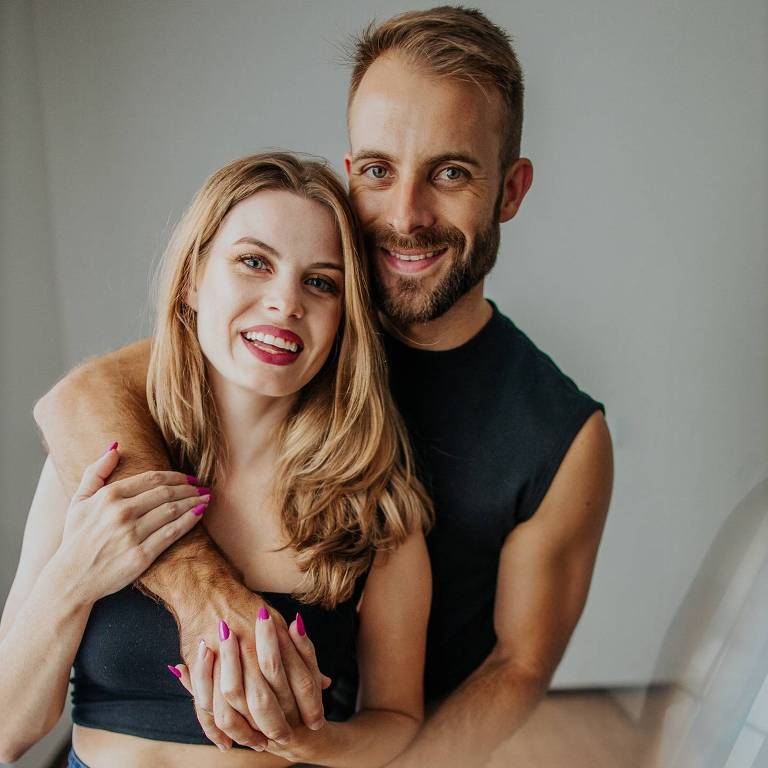 Um casal está posando para a foto. A mulher tem cabelo longo e liso, usa uma blusa preta e jeans. O homem está atrás dela, com um sorriso, usando uma camiseta preta. Eles estão se abraçando e parecem felizes. O fundo é neutro e claro, com pouca decoração visível.