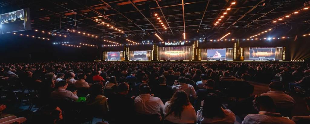 A imagem mostra um grande auditório lotado com uma multidão de pessoas assistindo a uma apresentação no palco. O palco é iluminado e possui telas grandes exibindo conteúdo. O ambiente é escuro, com luzes brilhantes penduradas no teto, criando uma atmosfera vibrante. As pessoas estão sentadas em cadeiras, algumas com roupas formais, e a maioria está voltada para o palco.
