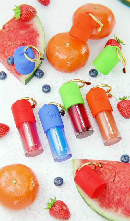 A imagem apresenta vários frascos de produtos de beleza em cores vibrantes, dispostos sobre uma superfície branca. Os frascos têm tampas coloridas em vermelho, azul, verde e laranja. Ao redor dos frascos, há frutas como melancia, tangerina, morangos e mirtilos, criando uma composição alegre e fresca.