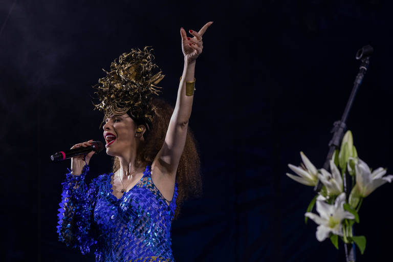 Uma artista se apresenta em um palco, usando um vestido azul brilhante e um adereço de cabelo dourado. Ela está segurando um microfone com a mão esquerda e levantando a mão direita em um gesto de empolgação. Ao lado dela, há um microfone em um suporte e algumas flores brancas visíveis no canto da imagem.