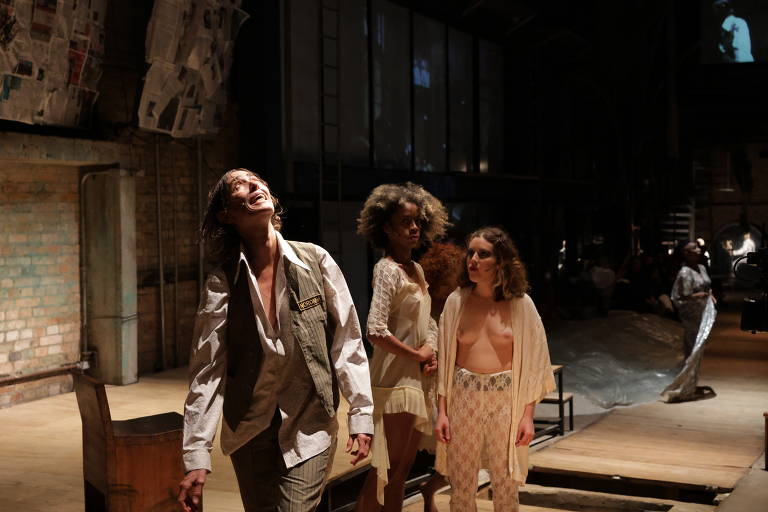 A imagem mostra uma cena de uma performance teatral em um espaço amplo e industrial. No centro, um homem com cabelo longo e solto, vestindo um colete e uma camisa branca, está sorrindo e olhando para cima. Ao seu lado, duas mulheres estão em trajes leves; uma delas tem cabelo cacheado e a outra tem cabelo liso e solto. Ao fundo, há uma projeção na parede e algumas pessoas assistindo à performance. O ambiente é iluminado de forma dramática, com paredes de tijolos expostos e um piso de madeira.