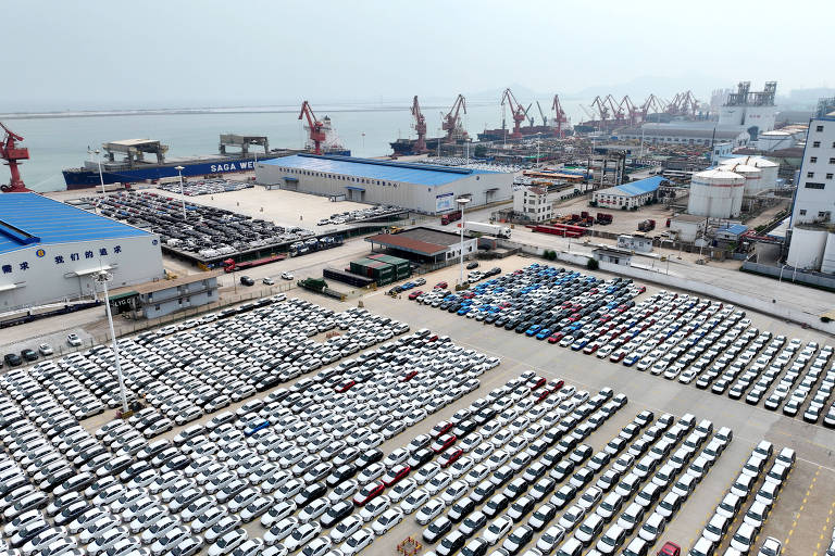 A imagem mostra uma grande área de estacionamento com muitos carros, predominantemente brancos, organizados em filas. Ao fundo, há um porto com guindastes e navios, além de edifícios industriais. O céu está nublado, indicando uma atmosfera de poluição ou neblina.