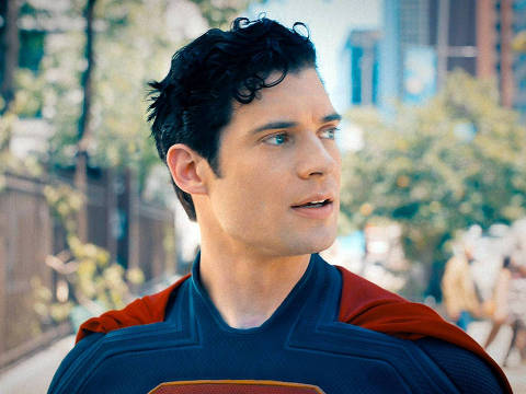 Cena do filme  Superman com David Corenswet  *** Local Caption *** A imagem mostra um jovem homem com cabelo cacheado e escuro, vestindo um traje de super-herói azul com uma capa vermelha. Ele está em um ambiente urbano, com árvores e edifícios ao fundo. O homem parece estar olhando para o lado, com uma expressão de curiosidade ou atenção.