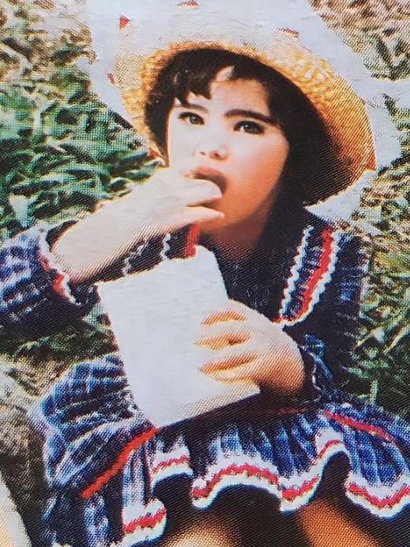 Uma criança sentada em um ambiente natural, usando um vestido listrado em azul e vermelho e um chapéu de palha. Ela está segurando um recipiente branco e levando a mão à boca, como se estivesse comendo algo. Ao fundo, há grama e vegetação.