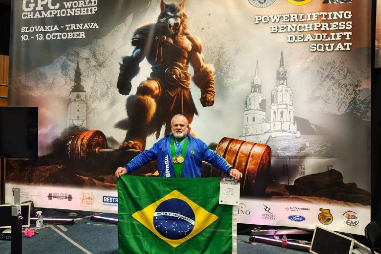 Um homem de cabelos grisalhos e barba, vestindo uma jaqueta azul com a palavra 'Brasil' e medalhas ao redor do pescoço, segura uma bandeira do Brasil em frente a um fundo que exibe um lobo antropomórfico e elementos relacionados ao powerlifting, como pesos e um certificado.