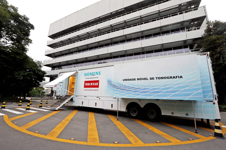 A imagem mostra uma unidade móvel de tomografia estacionada em frente a um edifício de vários andares. O veículo é da Siemens e possui uma escada de acesso. O fundo é composto por árvores e um edifício com varandas. A área ao redor do veículo tem faixas amarelas e cones de sinalização.