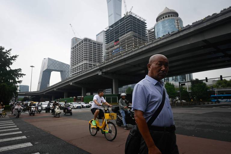A imagem mostra um cenário urbano com prédios em construção e um viaduto. No primeiro plano, um homem caminha em direção à câmera, enquanto um ciclista passa ao seu lado em uma bicicleta amarela. Ao fundo, há vários edifícios modernos e em construção, com guindastes visíveis. O céu está nublado, sugerindo um dia cinzento.