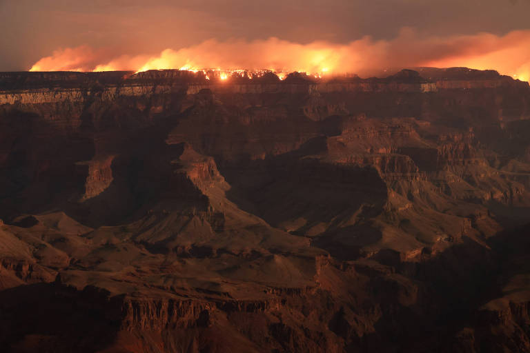 A imagem mostra uma vista do Grand Canyon ao entardecer, com fumaça e chamas visíveis no topo das montanhas. O céu está iluminado por um brilho laranja, indicando a presença de fogo. As formações rochosas do canyon são visíveis em primeiro plano, com sombras profundas e detalhes das camadas de rocha.