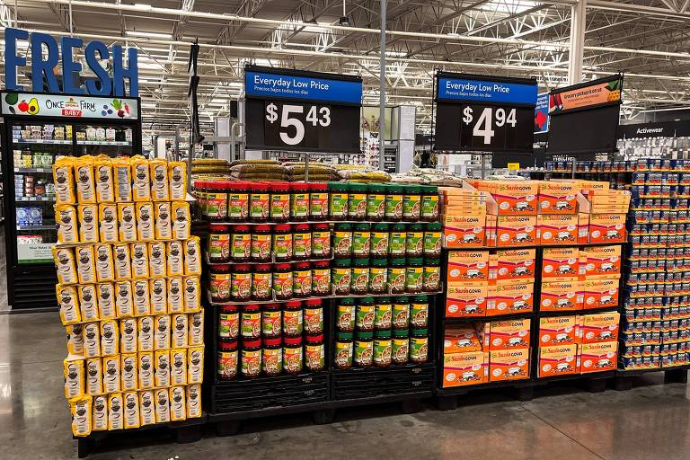A imagem mostra uma seção de supermercado com prateleiras organizadas. À esquerda, há várias caixas de produtos amarelos, possivelmente grãos ou cereais, com o preço de $5,43. No centro, há frascos de molho ou conservas em várias cores, e à direita, caixas de produtos laranja, com o preço de $4,94. Acima das prateleiras, há placas indicando os preços e a seção é bem iluminada.