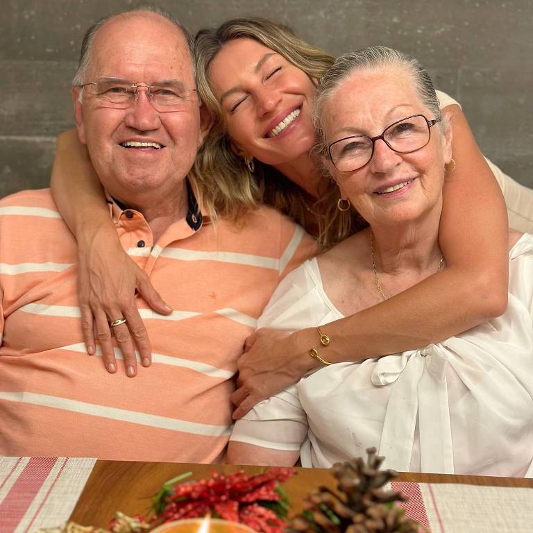 A imagem mostra três pessoas sorrindo em uma mesa decorada. Um homem idoso, à esquerda, usa uma camisa listrada em tons de laranja e branco. No centro, uma mulher jovem está abraçando o homem, sorrindo amplamente. À direita, uma mulher idosa usa óculos e uma blusa branca, também sorrindo. A mesa tem uma decoração de Natal com pinhas, folhas verdes e uma vela acesa.