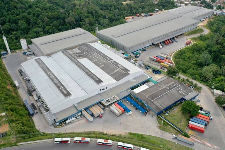 The image shows an aerial view of a large, flat-roofed warehouse located in a green area. The warehouse is light-colored and has several loading docks. Surrounding it are parked trucks and a curved road at the bottom of the image. In the background, an urban area with houses and vegetation can be seen.