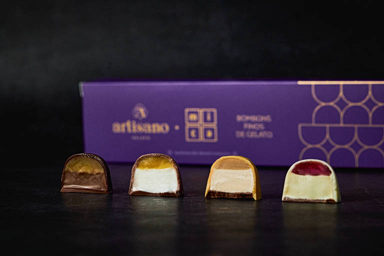 A imagem mostra uma caixa de bombons de gelato da marca Artisano, com quatro bombons dispostos em linha. Cada bombom tem uma camada de chocolate e diferentes recheios visíveis, que variam em cores e texturas. A caixa é de cor roxa com detalhes em dourado, contendo o nome da marca e a descrição dos produtos.