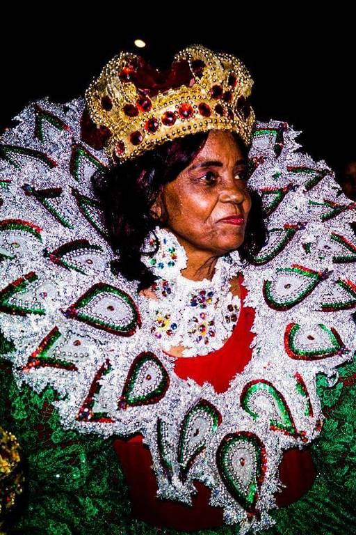 A imagem mostra uma mulher vestida com um traje tradicional exuberante, que inclui uma coroa dourada e um colar decorado. O traje é adornado com elementos verdes e brancos, criando um efeito visual impressionante. O fundo é escuro, destacando a figura da mulher e seu traje colorido.