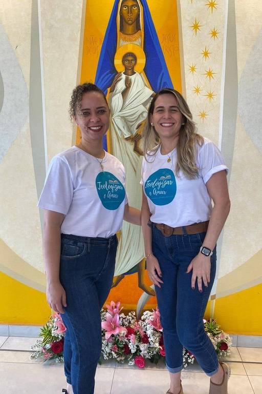 Deux femmes posent devant une image religieuse. Elles portent toutes deux des t-shirts blancs ornés d'un cercle bleu au centre, sur lequel est inscrit un texte. Elles sourient et portent un jean. En arrière-plan, on voit l'image d'une figure religieuse, sur fond jaune avec des détails blancs et dorés. Au sol, des fleurs décoratives.