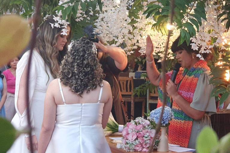 L'image montre une cérémonie de mariage en plein air, avec des fleurs blanches suspendues en arrière-plan. Deux mariées sont debout, l'une aux cheveux longs et détachés, l'autre aux cheveux bouclés. Un célébrant, vêtu d'une robe colorée, se tient devant elles et fait un geste de bénédiction. Une composition florale est posée sur une table à proximité, et quelques personnes sont visibles à l'arrière-plan, observant la cérémonie.