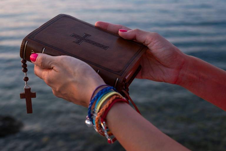 Une personne tient une Bible à couverture brune, ornée d'une croix. Un chapelet y est accroché, et la personne porte plusieurs bracelets colorés au poignet. L'arrière-plan, où se reflète une douce lumière, suggère que la scène se déroule au bord d'un lac ou de la mer.