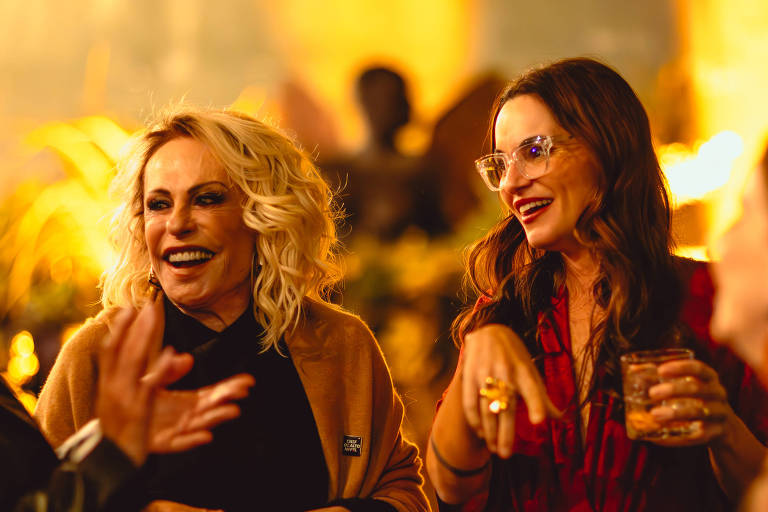 Um grupo de mulheres conversando em um ambiente iluminado, com um fundo desfocado que sugere uma festa ou evento social. Algumas mulheres estão sorrindo e gesticulando, enquanto outras seguram copos. As roupas variam em estilo, com uma mulher usando um vestido vermelho e outra com cabelo loiro e ondulado. O ambiente é acolhedor e festivo, com luzes quentes ao fundo.