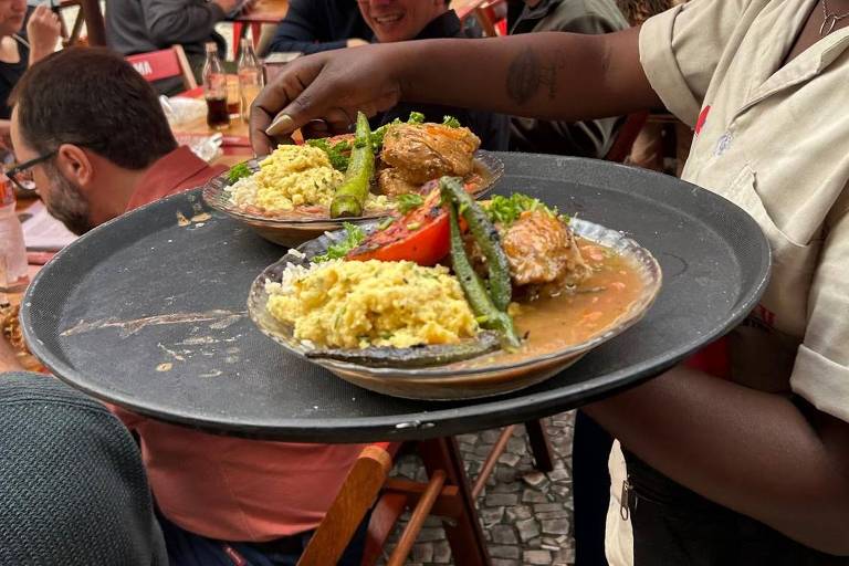 Uma garçonete está servindo dois pratos em uma bandeja. Os pratos contêm arroz, carne e vegetais, como pimentões e aspargos. O ambiente é de um restaurante ao ar livre, com mesas e cadeiras de madeira, e várias pessoas estão sentadas ao fundo, algumas conversando e outras olhando para a garçonete. Há guarda-sóis vermelhos cobrindo as mesas.