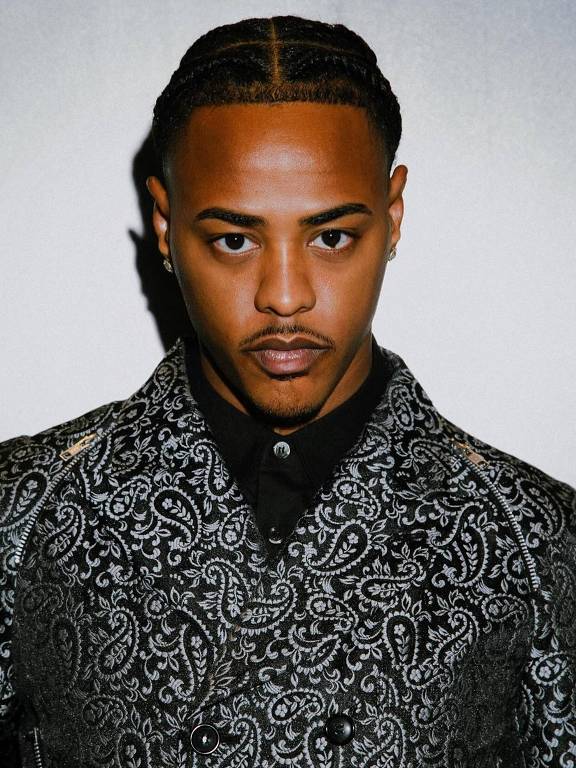Um homem posando para a câmera, vestindo uma camisa de padrão paisley em preto e branco. Ele tem cabelo trançado e uma expressão séria. O fundo é neutro, destacando sua roupa e aparência.