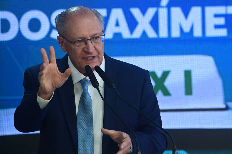 Um homem de terno e gravata, com cabelo grisalho, está falando em um microfone. Ele gesticula com a mão direita enquanto segura o microfone com a mão esquerda. Ao fundo, há uma tela com a palavra 'DOSTAXÍMETRO' visível. O ambiente parece ser um evento formal.